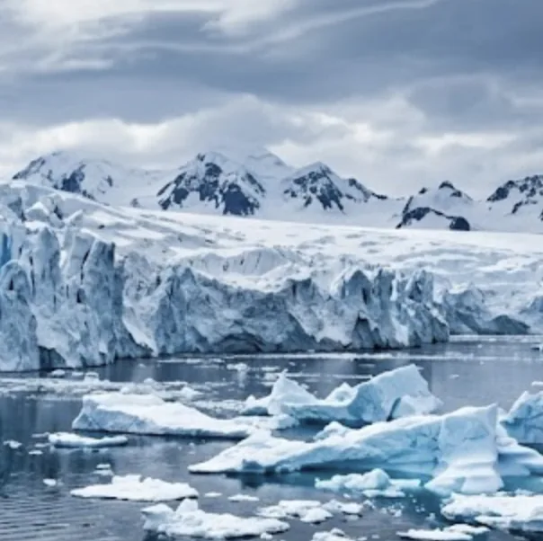 A tidewater glacier meeting the sea beneath snow-capped mountains, with broken ice floating in the foreground.